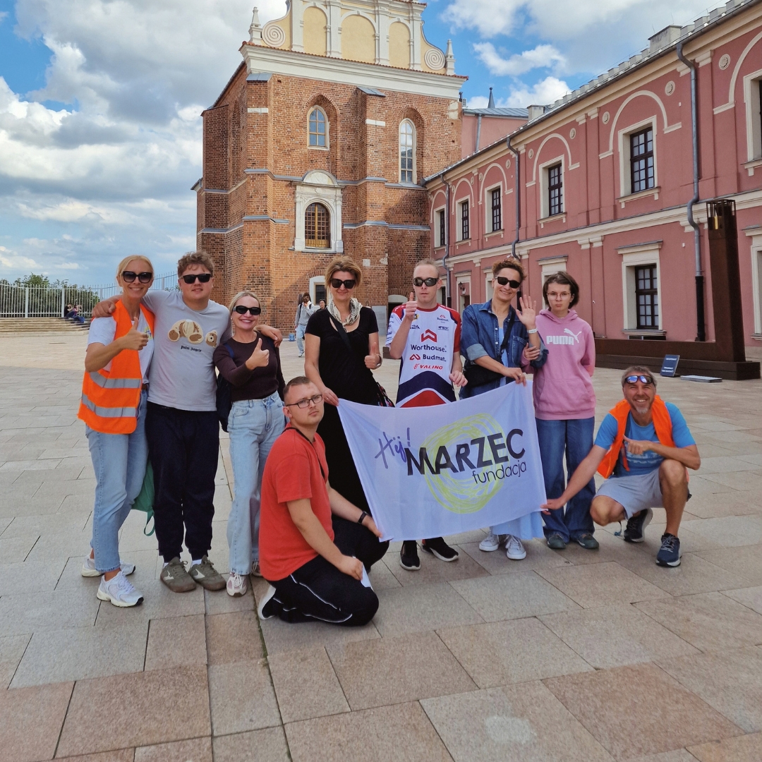 Grupa osób z flagą Fundacji Hej! MARZEC przed zabytkowym budynkiem w Lublinie.